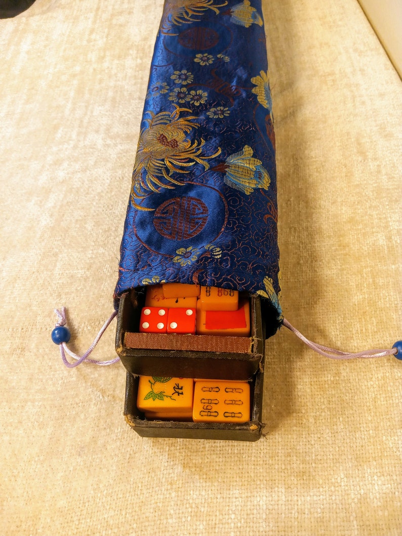 May include: A vintage Mahjong set with orange tiles and red dice, stored in a blue silk pouch with floral and circular patterns. The pouch has a drawstring closure with blue beads.