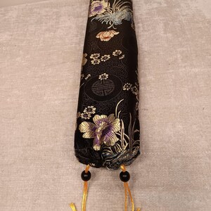 May include: A black, cylindrical fabric case with a floral pattern in gold, purple, and white. The case has a drawstring closure with black beads and gold cords. The floral design is set against a black background.