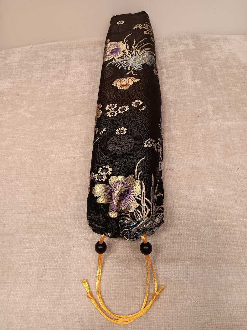 May include: A black, cylindrical fabric case with a floral pattern in gold, purple, and white. The case has a drawstring closure with black beads and gold cords. The floral design is set against a black background.