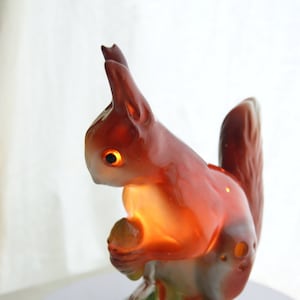 Puede incluir: Lámpara de cerámica con forma de ardilla en tonos marrón y naranja, sosteniendo una nuez. La lámpara tiene ojos brillantes y una luz cálida y acogedora. La base es blanca y la lámpara está enchufada.