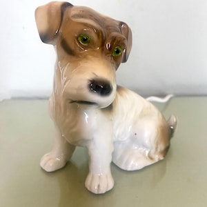 Könnte beinhalten: Eine Keramikfigur eines sitzenden Hundes. Der Hund hat ein braun-weißes Fell, grüne Augen und eine schwarze Nase. Die detaillierte Figur scheint einen Jack Russell Terrier darzustellen.