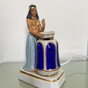 Könnte beinhalten: Eine ägyptisch thematisierte Keramiklampe mit einer Figur einer Frau in traditioneller Kleidung. Die Lampe hat einen blau-weißen Sockel mit goldenen Akzenten. Die Frau hält eine Schale, und die Lampe hat ein dekoratives Design.