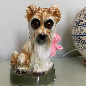 Peut inclure: Figurine de chien en céramique avec fourrure marron et blanche, grands yeux bruns et un nœud rose. Le chien est assis sur une base verte. Un objet décoratif en céramique est en arrière-plan.