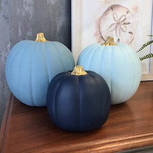 May include: Three decorative pumpkins in varying shades of blue, with gold-colored stems. The pumpkins are arranged on a wooden surface, with a framed picture in the background. The pumpkins are a light blue, medium blue, and dark blue.
