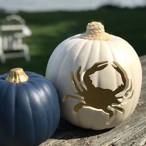 May include: Two decorative pumpkins. One is navy blue with a gold stem. The other is white with a crab cut-out design and a rope stem. The pumpkins are sitting on a wooden surface.