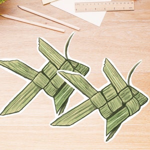 May include: Two green fish-shaped woven palm leaf decorations with white borders. The fish are detailed with pencil shading, and are placed on a light wood grain surface. The fish are a craft or decorative item.