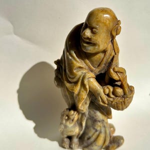 Peut inclure: Statuette en pierre sculptée d'un homme agenouillé, tenant un panier de fruits. La figurine est dans les tons de beige et de marron, avec un petit animal à ses pieds. La sculpture a une finition lisse et polie, sur fond blanc.