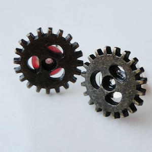 May include: A pair of bronze-colored metal earrings shaped like gears. The gears have a red accent in the center.