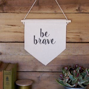 K&ouml;nnte beinhalten: Ein wei&szlig;es Canvas-Banner mit schwarzem Text, der "be brave" lautet, h&auml;ngt an einer Holzleiste an einer Holzwand. Das Banner h&auml;ngt an einer Schnur. Unter dem Banner befinden sich auf einer hellblauen Holzoberfl&auml;che B&uuml;cher, eine Garnrolle und eine Topfpflanze.