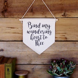 Könnte beinhalten: Ein kleines Holzbanner mit dem Text "Bind my wandering heart to Thee" hängt an einer rustikalen Holzwand. Das Banner ist weiß mit schwarzen Buchstaben und hängt an einer Schnur.