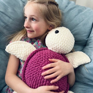 Puede incluir: Una niña pequeña está sentada en un sofá y sostiene un juguete de peluche de tortuga tejido a crochet. La tortuga es blanca con un caparazón rosa y ojos negros. La niña lleva un vestido floral y pantalones vaqueros azules.