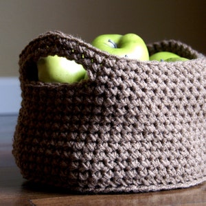 May include: A brown crocheted basket with a handle, filled with green apples.