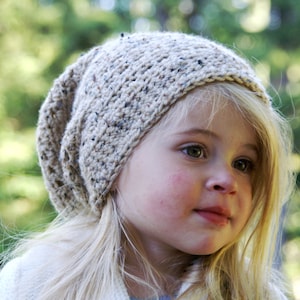 May include: A young girl with blonde hair wearing a beige knitted beanie with a flecked pattern.