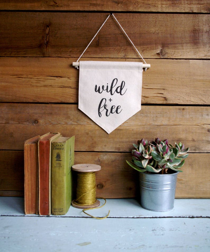 May include: A white canvas banner with the text "wild + free" hanging on a wooden wall. The banner is attached to a wooden dowel rod with twine. There are books, a spool of twine, and a potted succulent on a light blue shelf below the banner.