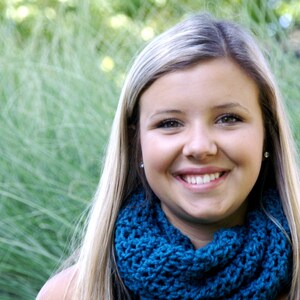 May include: A young woman with long blonde hair is wearing a teal blue crocheted infinity scarf. She is smiling and looking at the camera.