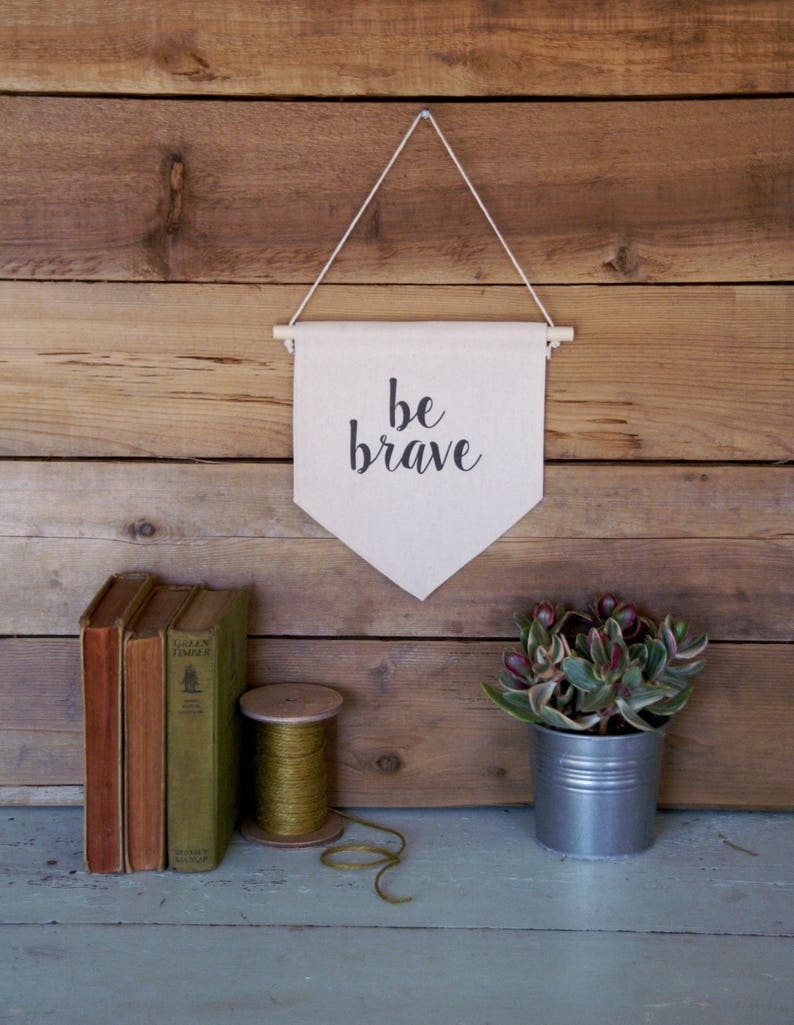 K&ouml;nnte beinhalten: Ein wei&szlig;es Canvas-Banner mit schwarzem Text, der "be brave" lautet, h&auml;ngt an einer Holzleiste an einer Holzwand. Das Banner h&auml;ngt an einer Schnur. Unter dem Banner befinden sich auf einer hellblauen Holzoberfl&auml;che B&uuml;cher, eine Garnrolle und eine Topfpflanze.