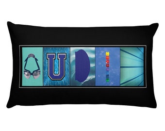 May include: A black rectangular pillow with a white border and a colorful design featuring the letters "U D I" with images of swimming goggles, a swimming pool, a kickboard, and a lane line.