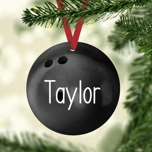 May include: A round, black bowling ball ornament featuring three finger holes and the name Taylor printed in white lettering. The ornament is suspended by a red ribbon from a green pine branch, creating a festive holiday decoration.