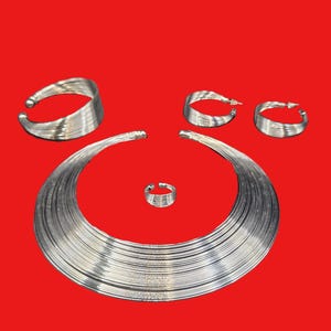 Puede incluir: Un conjunto de joyas plateadas sobre un fondo rojo. El conjunto incluye un collar ancho, una pulsera, un anillo y dos pares de pendientes de aro. Las joyas parecen estar hechas de múltiples hilos de alambre de metal.