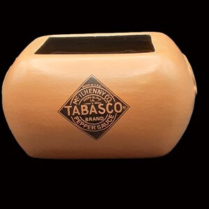 Könnte beinhalten: Ein terracottafarbener Keramik-Pflanztopf in Form einer Tabasco-Sauce-Flasche. Der Topf hat ein schwarzes Innenleben und trägt das Tabasco-Markenlogo in Schwarzweiß.