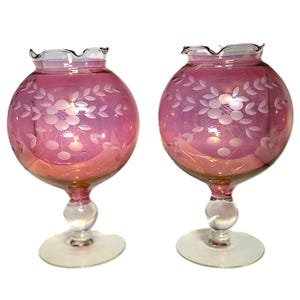 May include: Two pink glass candle holders with etched floral designs. The round bowls have a ruffled rim and sit atop a clear stem with a round ball and a circular base. The glass has a pink hue.