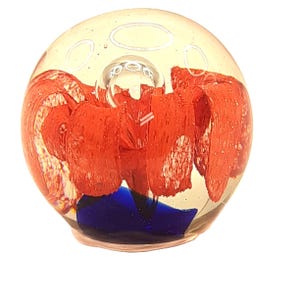 May include: A clear glass paperweight with red and blue abstract designs. The paperweight has a spherical shape with a large bubble in the center. The red design has a textured appearance. The base of the paperweight is a dark blue color.