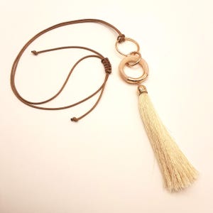 May include: A long necklace featuring a beige tassel pendant. The pendant is attached to a gold-colored ring with a beige center. The necklace has a brown cord with adjustable knots.