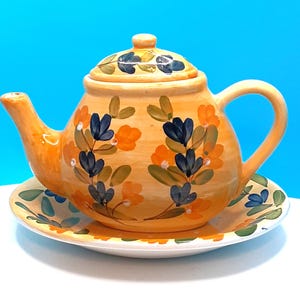 May include: A hand-painted ceramic teapot and saucer set. The teapot is a warm orange color, adorned with blue and orange floral designs. The matching saucer features similar floral patterns. The set is ideal for serving tea.