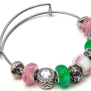 May include: Expandable silver bracelet with a selection of beads. The beads include pink, green, and white glass beads, plus silver and rhinestone details. The bracelet is designed to expand for easy wear.