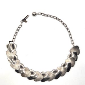 May include: A silver-toned necklace featuring a series of heart-shaped pendants. The pendants alternate between smooth and textured finishes, connected by a chain. The necklace has a clasp closure and a chain extender.