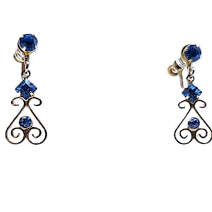 May include: A pair of vintage clip-on earrings. Each earring features a large, round, blue gemstone at the top, a square blue gemstone, and a smaller round blue gemstone. The earrings have a decorative, openwork design in a gold tone.