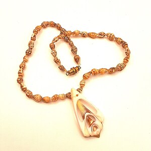 May include: A brown shell necklace with a large, white shell pendant. The pendant is carved with intricate details and has a gold-colored accent.