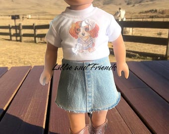 May include: A doll wearing a white t-shirt with a Cavalier King Charles Spaniel graphic, a denim skirt, and brown cowboy boots. The doll has blonde hair and is standing on a wooden surface. The text "Lillie and Friends" is on the shirt.
