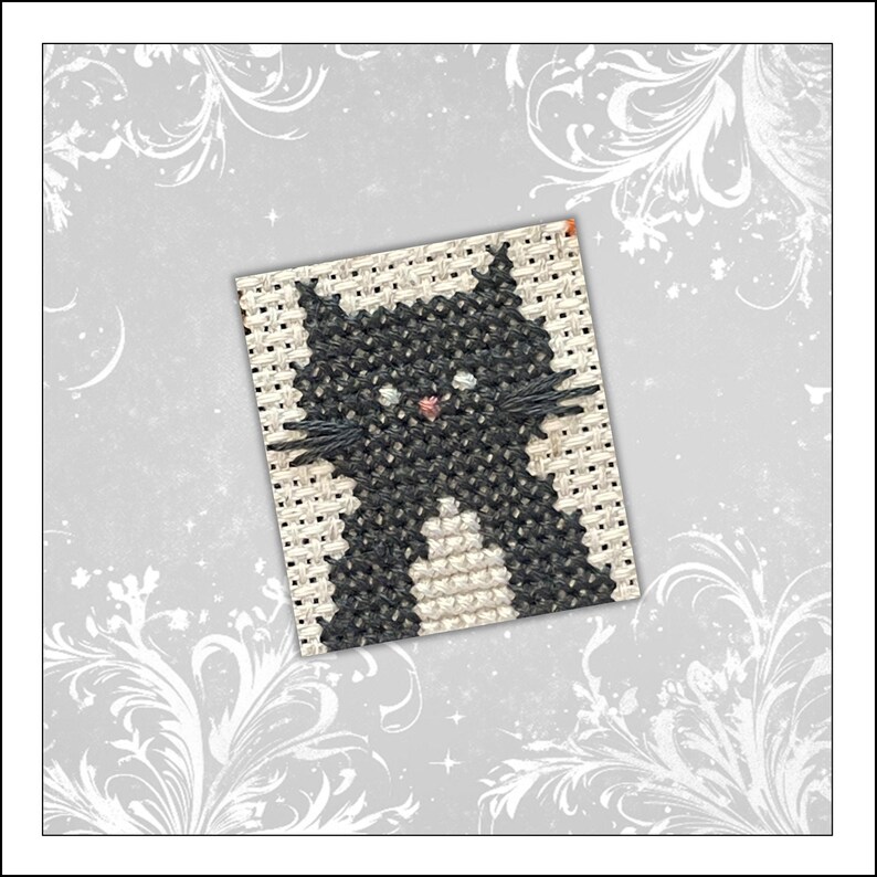 May include: A square cross-stitch embroidery piece on light beige fabric featuring a black cat with a white chest patch, small white eyes, and a tiny pink nose, accented by black thread whiskers extending from the sides of the cat's face.