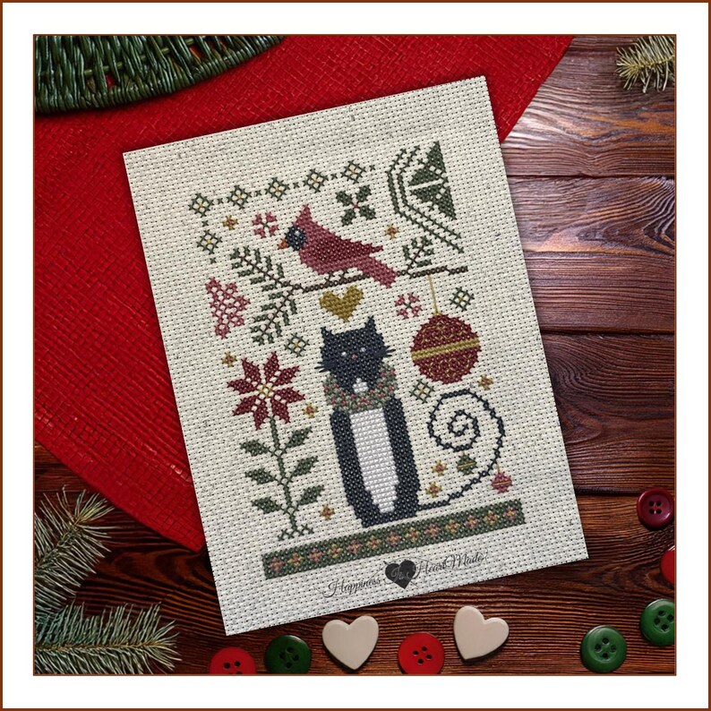 May include: A cross-stitch pattern on cream fabric featuring a black and white cat wearing a scarf, a red cardinal on a branch, a red ornament, and a poinsettia. The design includes geometric accents and the text Happiness Heart Made.