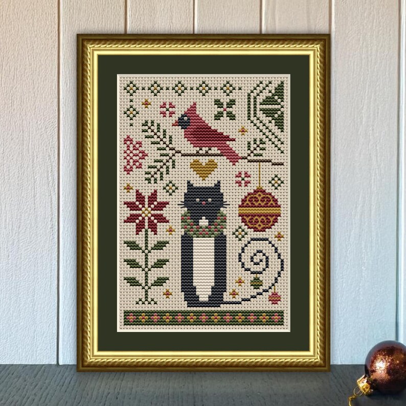May include: A framed cross-stitch embroidery featuring a black cat wearing a green and red scarf, positioned below a red cardinal on a branch, a gold heart, and a decorative ornament, all surrounded by floral and geometric patterns.