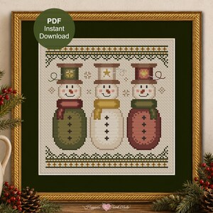 May include: A framed cross-stitch pattern featuring three snowmen. Each snowman has a different colored hat, scarf, and coat. The text "PDF Instant Download" is in a green circle in the upper left corner. The frame is gold.
