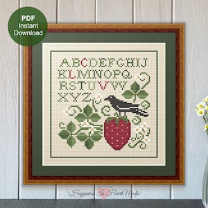May include: Cross stitch pattern featuring the alphabet, a bird, and a strawberry. The pattern is framed in a brown wood frame with a green mat. The text "Happiness Is Heart Made" is at the bottom of the frame.