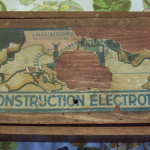 Può includere: Una scatola di legno con un'etichetta parzialmente strappata che recita "L'ELECTROTECHNIQUE" e "BOITE DE CONSTRUCTION ELECTROTECHNIQUE". La scatola è marrone e ha un aspetto usurato e vintage.