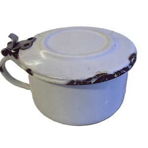 Könnte beinhalten: Weiße Emaille-Tasse mit Deckel und Henkel. Die Tasse hat eine abgerundete Form und einen Deckel, der gut aufsitzt. Die Ränder der Tasse und des Deckels haben eine verwitterte, braune Oberfläche, die ihr einen Vintage-Look verleiht. Der Henkel ist klein und gebogen.