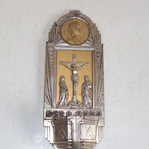 Peut inclure: Une décoration murale religieuse vintage représentant un crucifix avec Jésus sur la croix, flanqué de deux personnages. La pièce a une finition or et argent avec un design Art Déco et un petit portrait circulaire en haut.