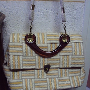 Peut inclure: Un sac à main tissé beige et blanc avec une poignée en plastique marron et des ferrures dorées. Le sac a une fermeture à rabat et un motif tissé.
