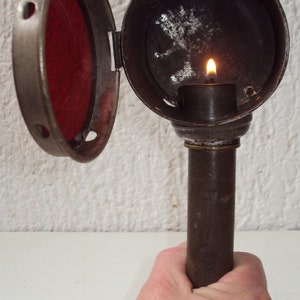 Puede incluir: Una linterna de vela vintage de metal con una lente de vidrio roja y una vela encendida en su interior. La linterna tiene un asa y una tapa con bisagras.