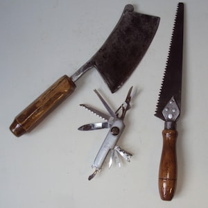 Peut inclure: Trois outils : un grand couperet en métal incurvé avec un manche en bois, un petit multi-outil argenté à lames multiples et une scie avec un manche en bois.