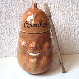 Peut inclure: Un pot à cornichons en céramique brune avec un visage souriant et le mot "Cornichons" inscrit sur le couvercle. Le pot est livré avec une cuillère en bois.