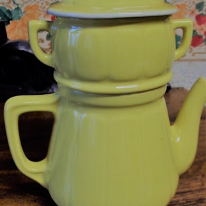 Puede incluir: Una tetera de cerámica amarilla con tapa y una olla separada para preparar café. La tetera tiene un diseño acanalado y un asa a cada lado.