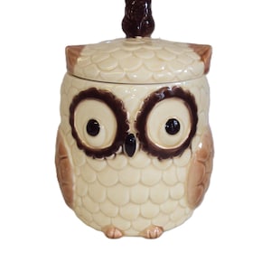 Peut inclure: Un pot à biscuits en céramique en forme de hibou. Le pot est de couleur crème avec des accents bruns, notamment les yeux, les oreilles et une petite pièce décorative sur le couvercle. Le corps du hibou est texturé pour ressembler à des plumes.