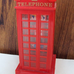 Könnte beinhalten: Eine rote Telefonzelle mit dem Wort "TELEPHONE" in goldener Schrift. Die Zelle hat einen rechteckigen Sockel und eine Tür mit mehreren Glasscheiben. Der Hintergrund ist eine Holzoberfläche.