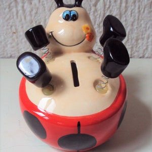 Peut inclure: Une tirelire en forme de coccinelle rouge et noire avec un visage blanc, des yeux noirs et une bouche noire. La coccinelle a des pattes à ressorts et une fente sur le dos pour les pièces de monnaie.