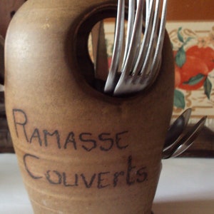 Peut inclure: Un porte-ustensiles en céramique marron avec une poignée et une grande ouverture. Des fourchettes et des cuillères en argent sont visibles. Les mots "RAMASSE COUVERTS" sont écrits sur le devant. Un article de cuisine décoratif.